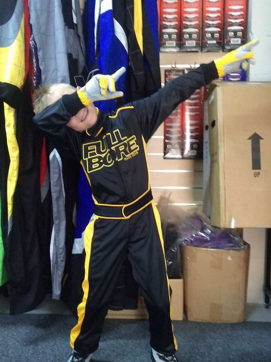 Child posing playfully in a black and yellow Full Bore racing suit and gloves inside a store with racing gear hanging on display and boxes in the background.